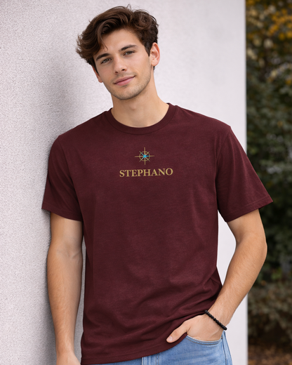"Jonge man draagt burgundy kleur T-shirt met gouden ‘STEPHANO’ logo en klein kompas symbool op de borst, gecombineerd met lichtblauwe jeans.