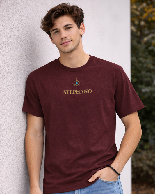 "Jonge man draagt burgundy kleur T-shirt met gouden ‘STEPHANO’ logo en klein kompas symbool op de borst, gecombineerd met lichtblauwe jeans.